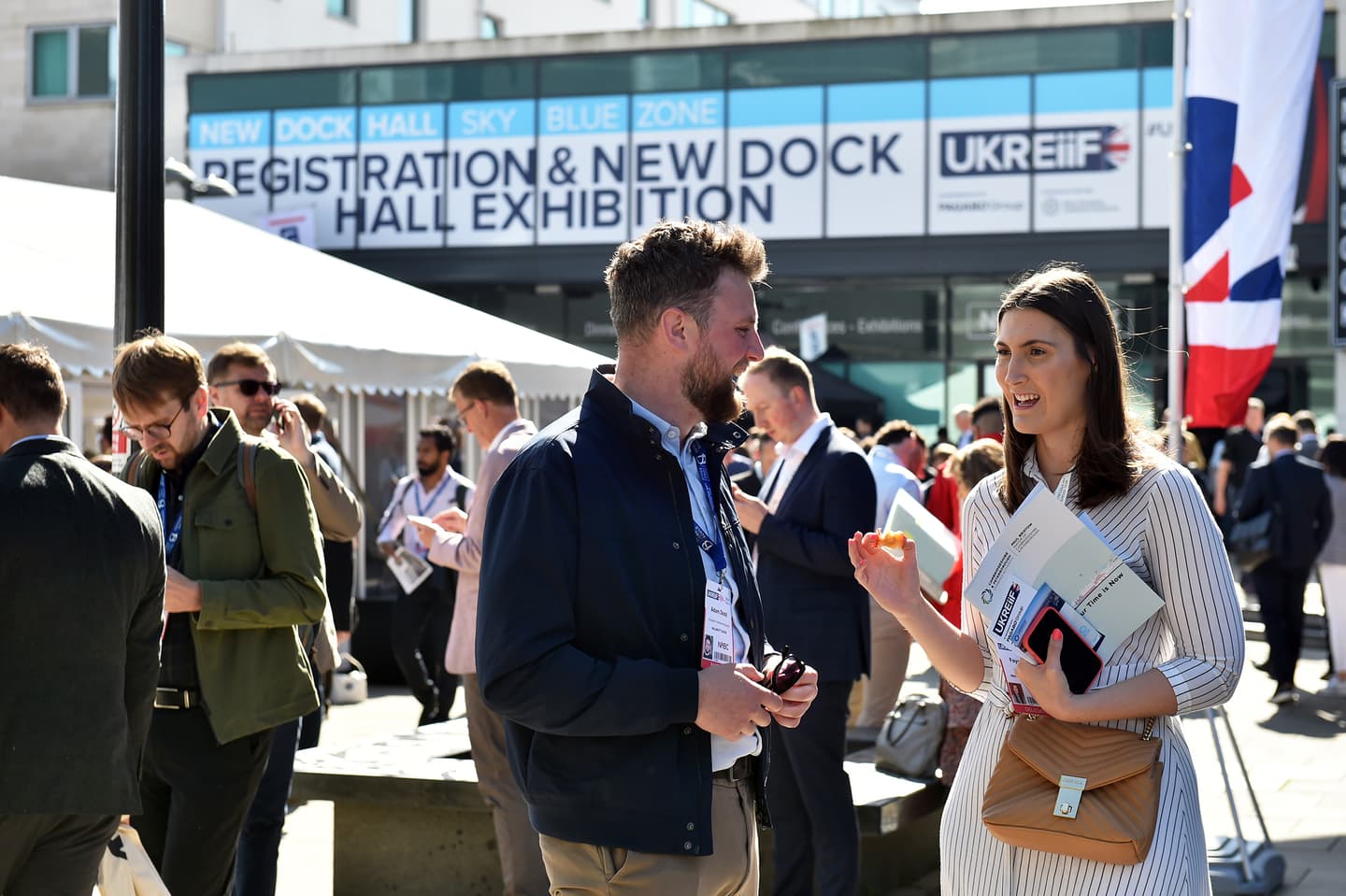 People stand and talk outside a building with a large sign reading Registration & New Dock Hall Exhibition during a business event.
