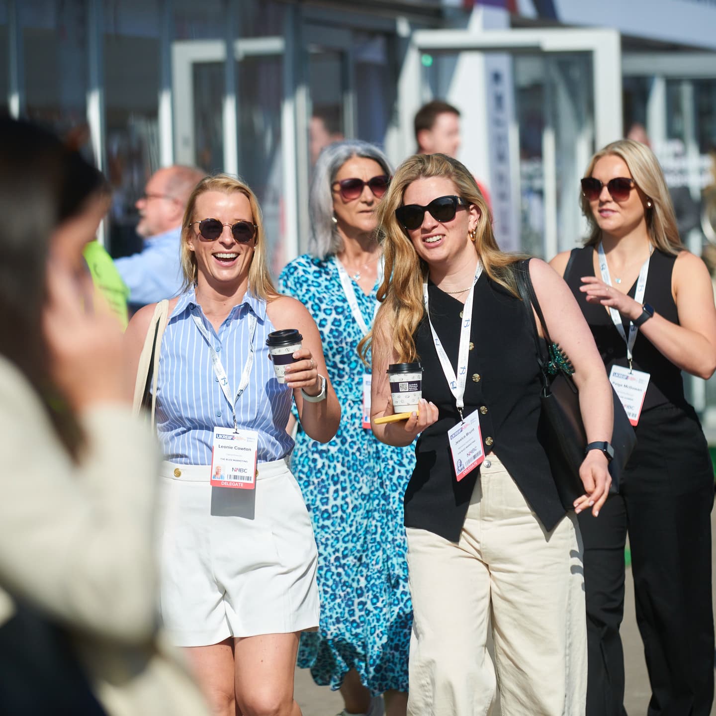 A group of women in business attire walk outdoors at an event, wearing name badges and sunglasses, whilst smiling and talking.