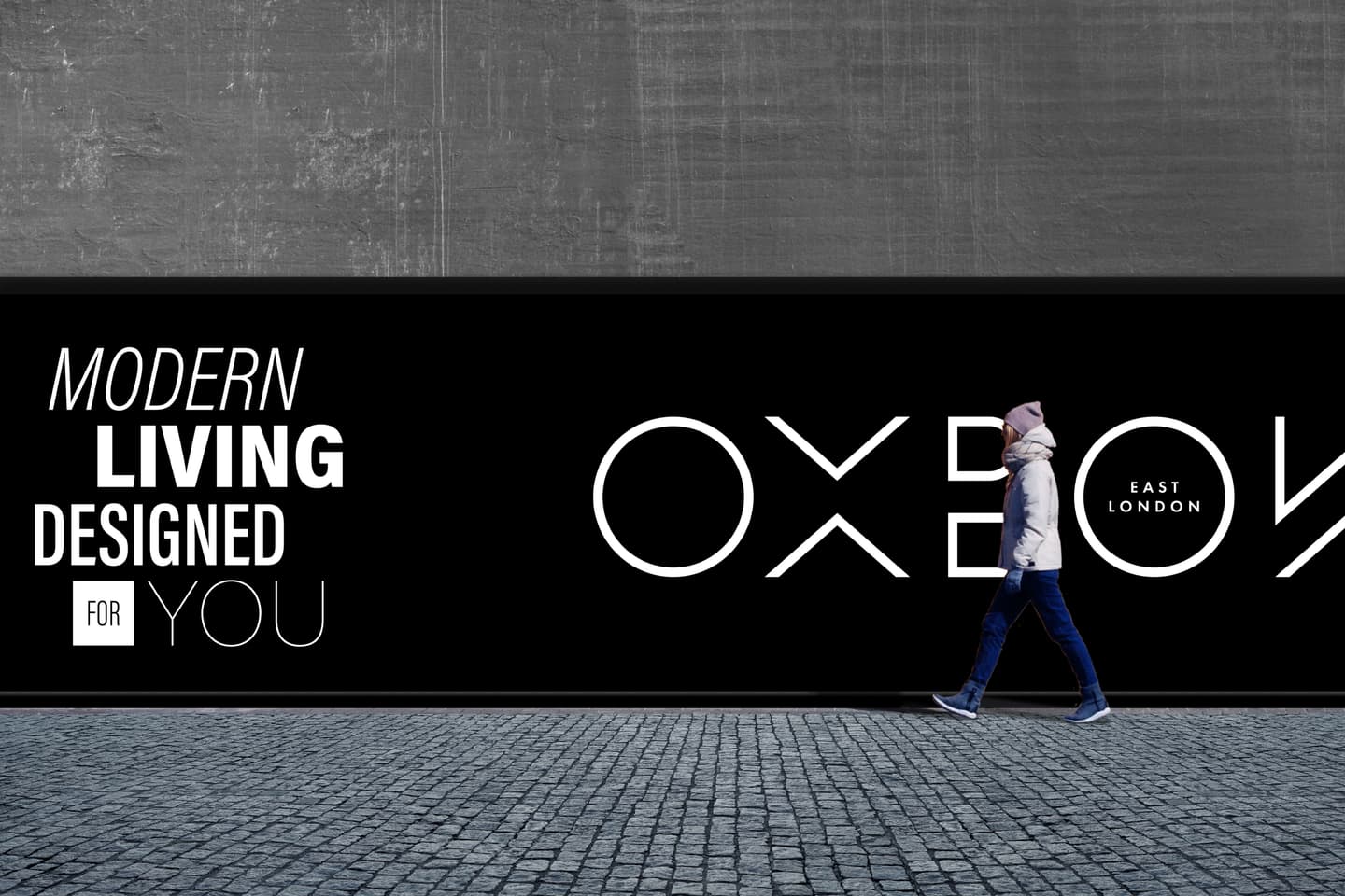 A person walks past a large black and white hoarding with the text Modern living designed for you and OXBOX East London on a cobbled pavement.