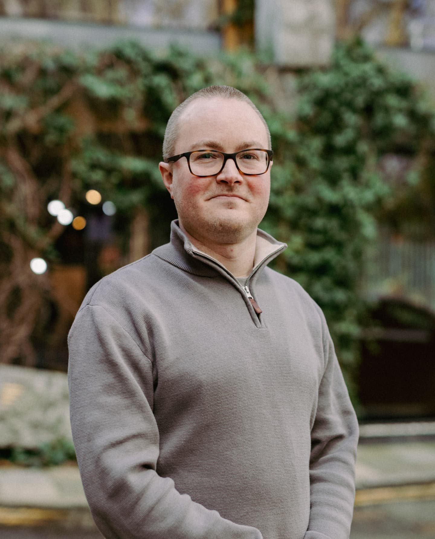 A man with glasses and short hair stands outdoors, wearing a light grey quarter-zip jumper, with greenery and a blurred building in the background.