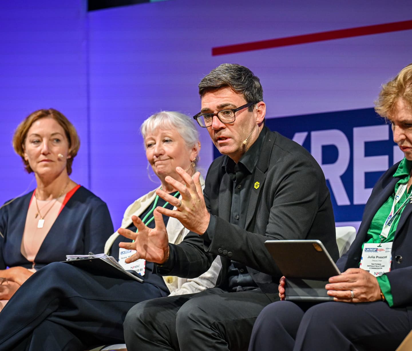 Four panellists sit on stage at a conference, one man speaking animatedly while the others listen, all wearing name badges and holding notes or tablets.