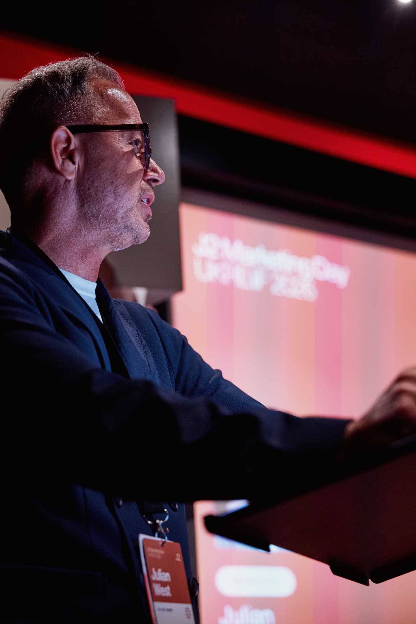 A man wearing glasses speaks at a lectern during a Marketing Day event; a presentation screen with event details is visible in the background.