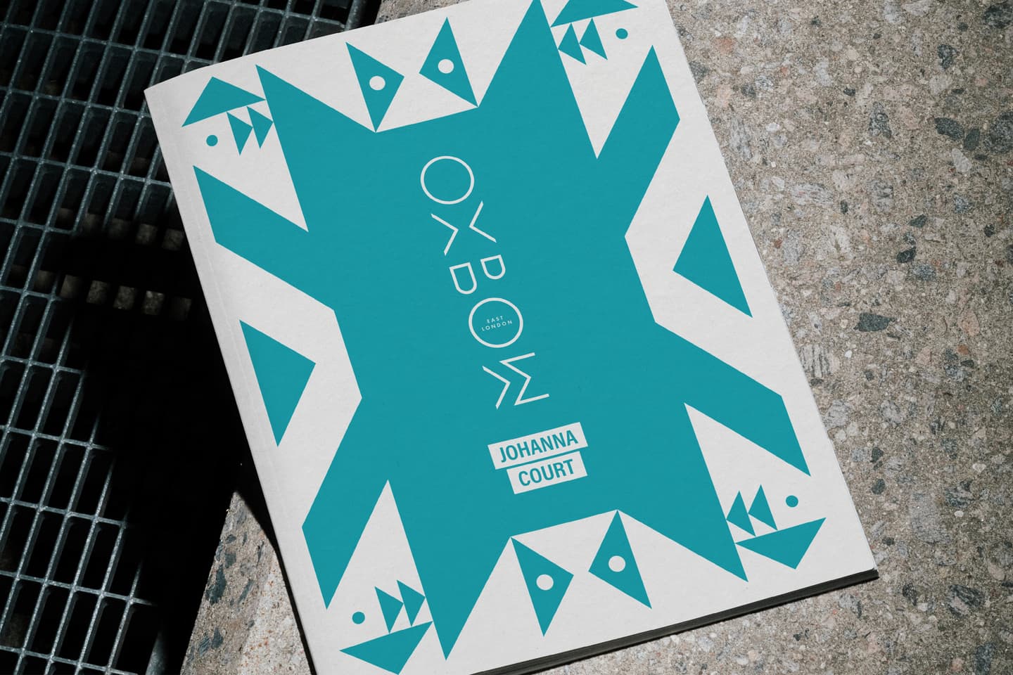 A booklet titled OXBOW by Johanna Court features a geometric teal design on a concrete surface next to a metal grating.