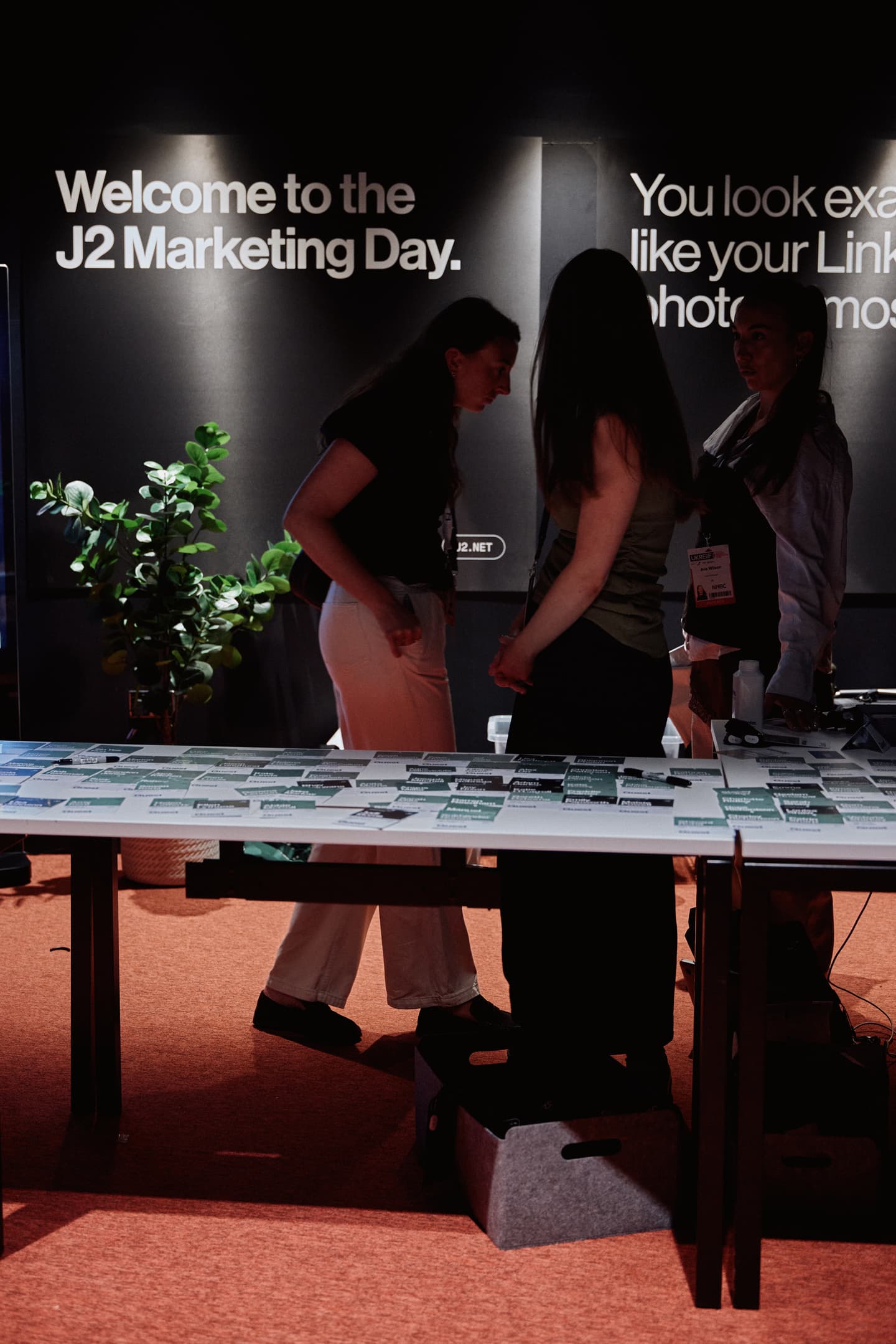 Three people stand near a table with name tags at an event with signs reading Welcome to the J2 Marketing Day. and You look exactly like your Link.