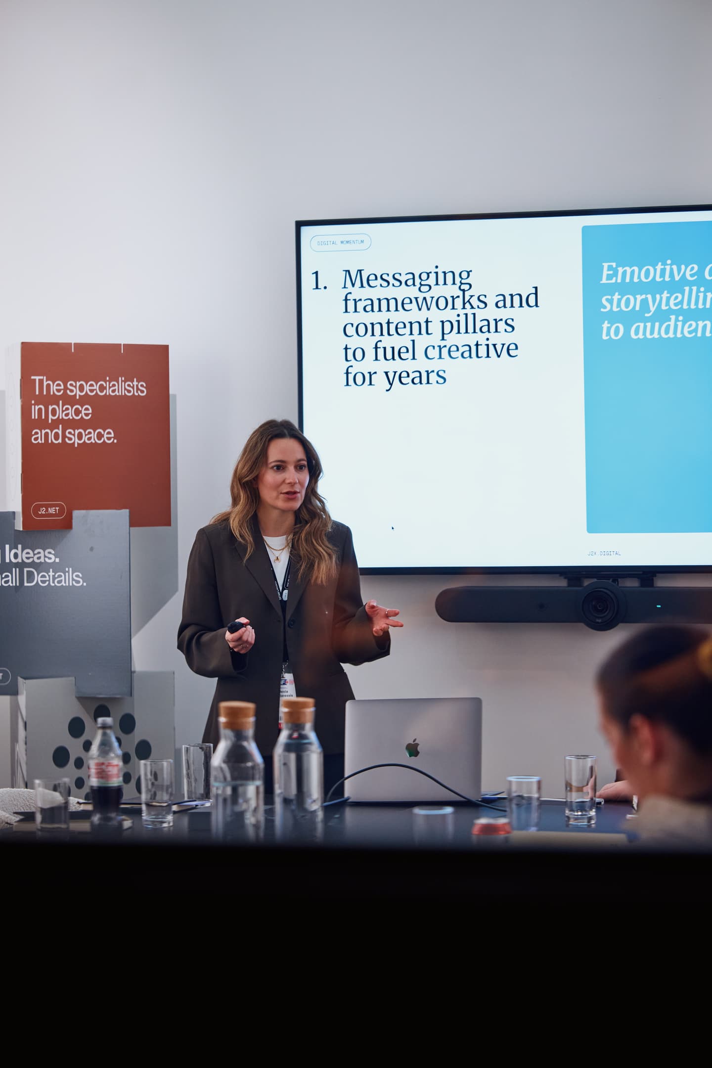 A woman stands and presents to a group; a screen behind her displays slides about messaging frameworks and storytelling. Bottles and laptops are visible on the table.