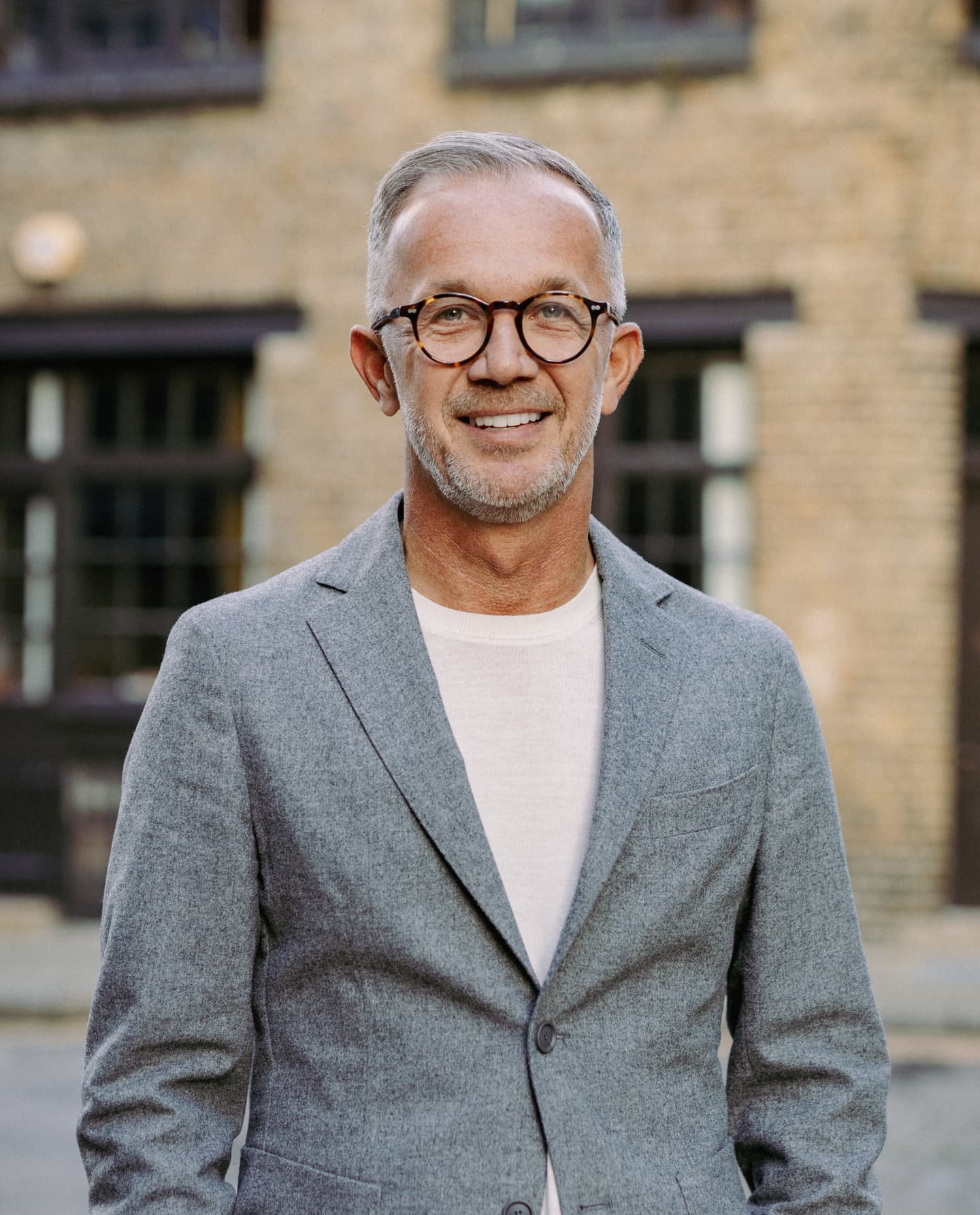 A man with short grey hair, glasses, and a beard is wearing a grey blazer over a white shirt, standing outdoors in front of a brick building.
