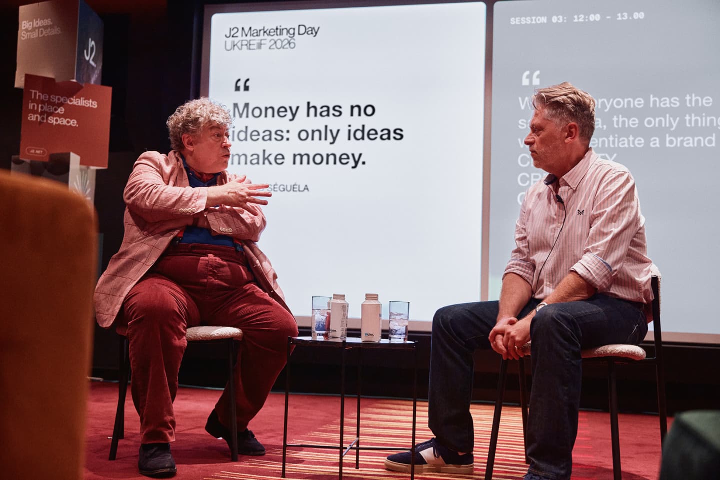 Two men sit and talk on stage at a marketing event, with a presentation slide behind them displaying a quotation about money and ideas.