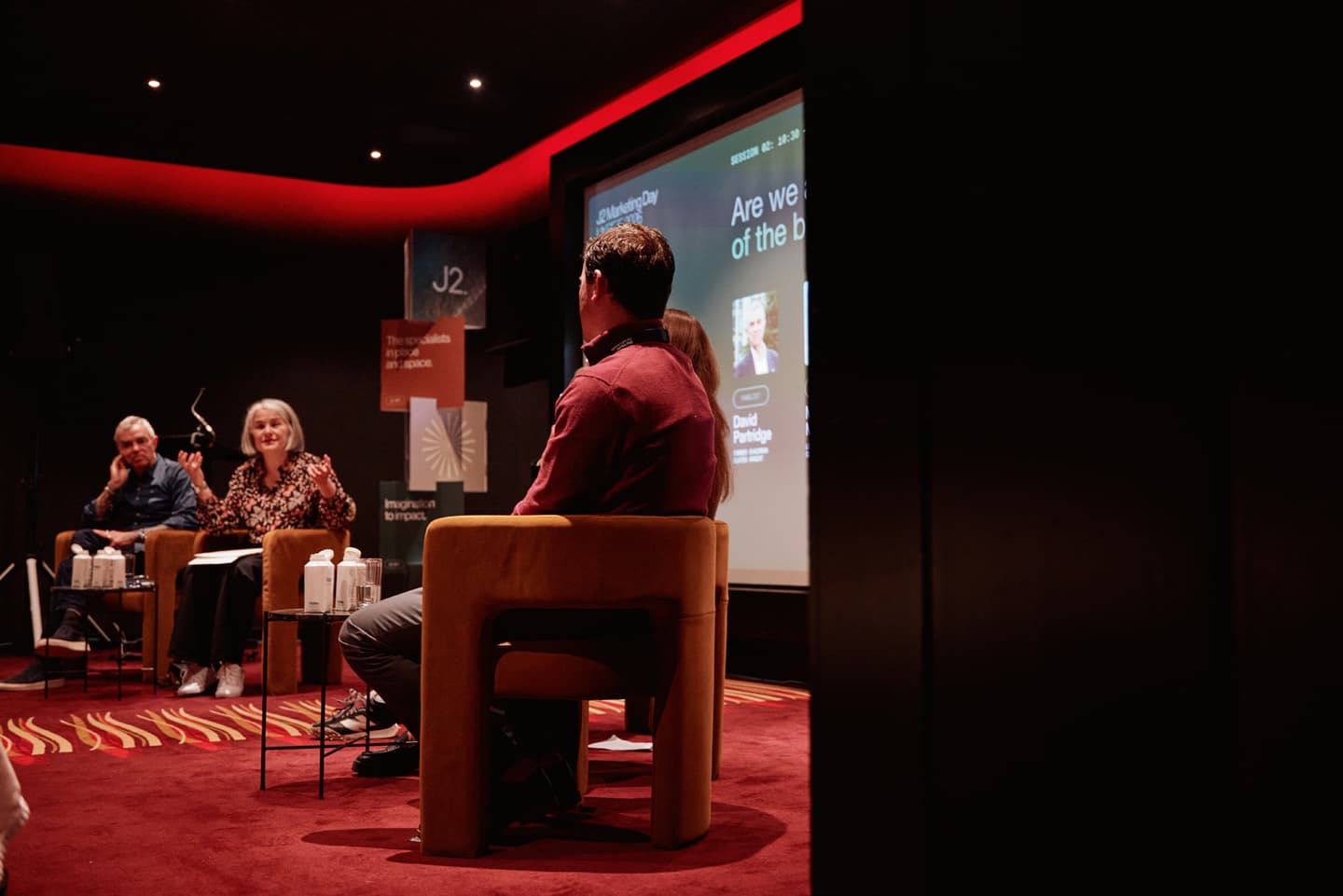 A panel of speakers sits on stage in a dimly lit auditorium, engaged in discussion with a projected presentation behind them.