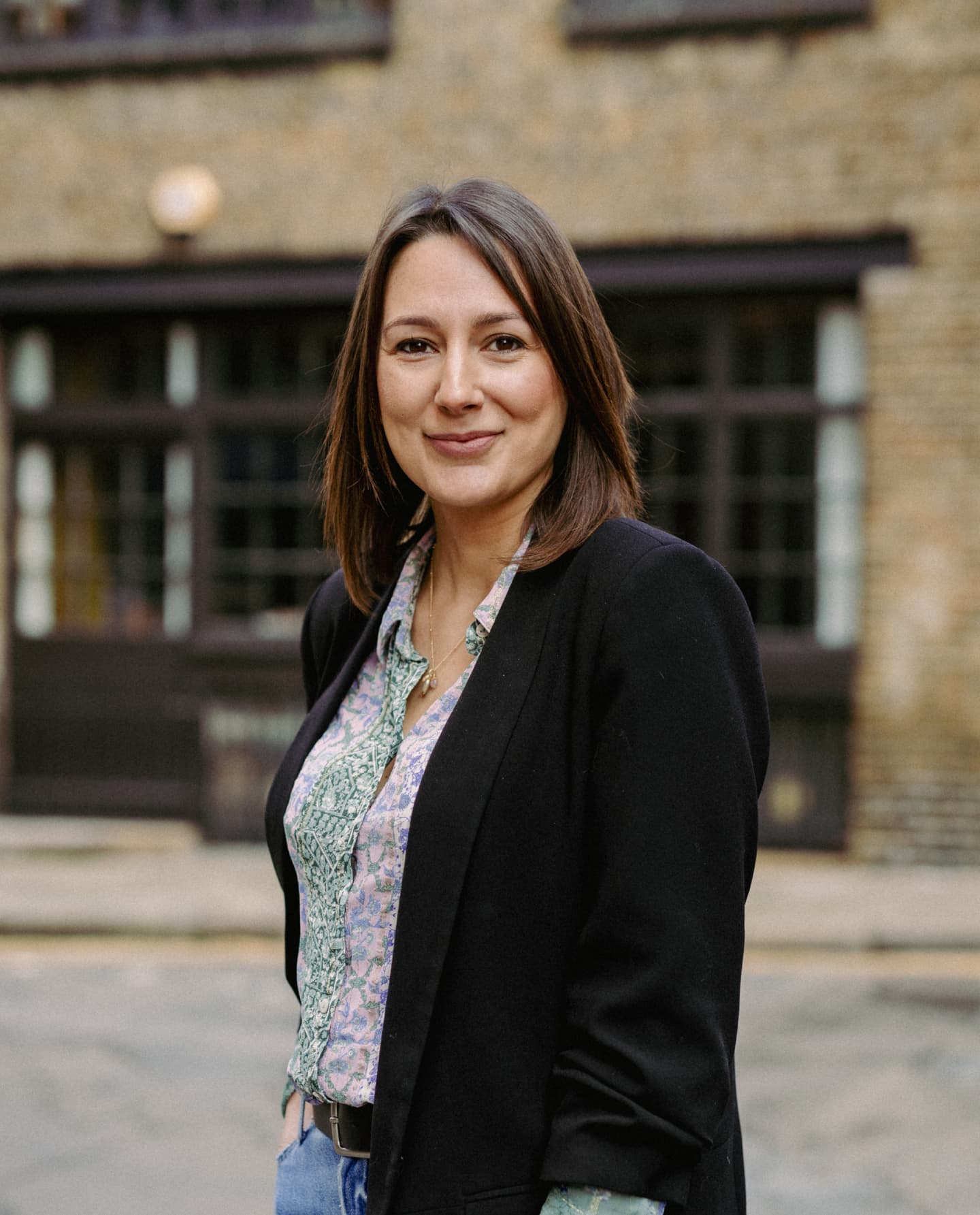 A woman with straight brown hair stands outdoors in front of a brick building, wearing a black blazer over a patterned shirt and blue jeans, smiling at the camera.