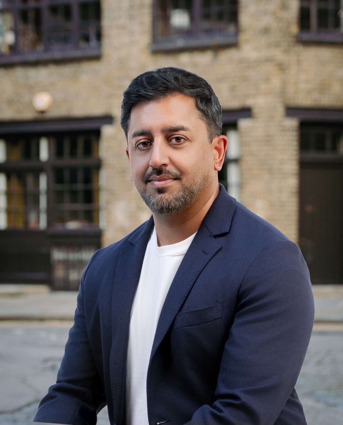 A man with short dark hair and a beard, wearing a navy blazer and white shirt, sits outdoors in front of a brick building.